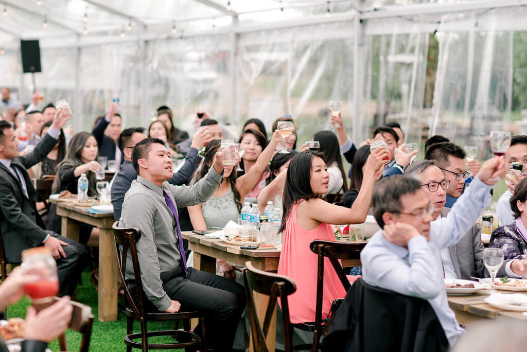 Beautiful outdoor reception in a clear tent surrounded by family and friends giving cheers. https://www.thegathered.ca