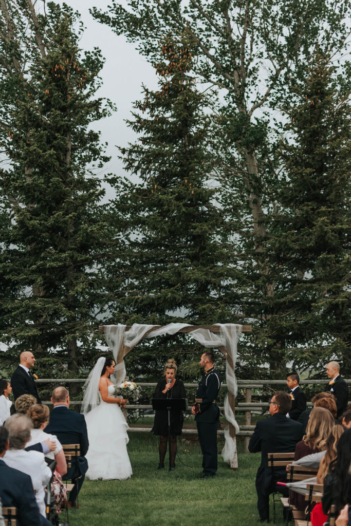 couple standing at the alter under a decorated wood arbor and big pine trees in the background
