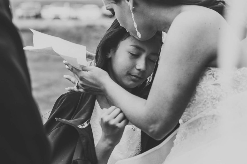 black and white image of bride hugging the flower girl while holding her vows in her hand