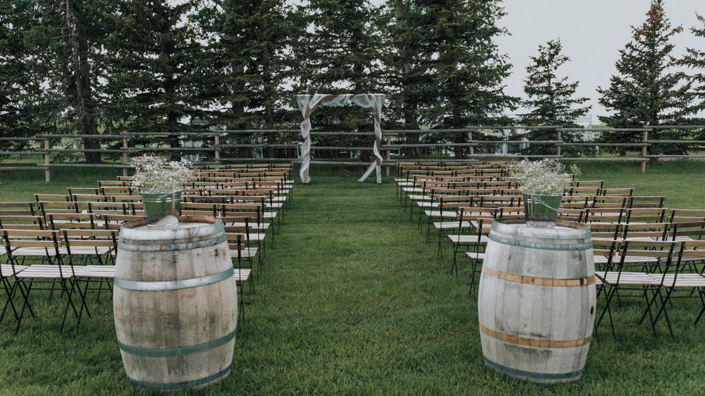 outdoor wedding ceremony space with trees in the background, wood arbor, wood slotted chairs and two wine barrels at the end of the aisle