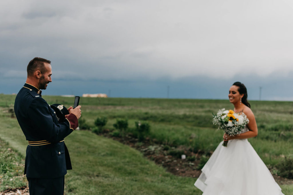 groom taking a picture of his bride with his phone in a green field with storm clouds