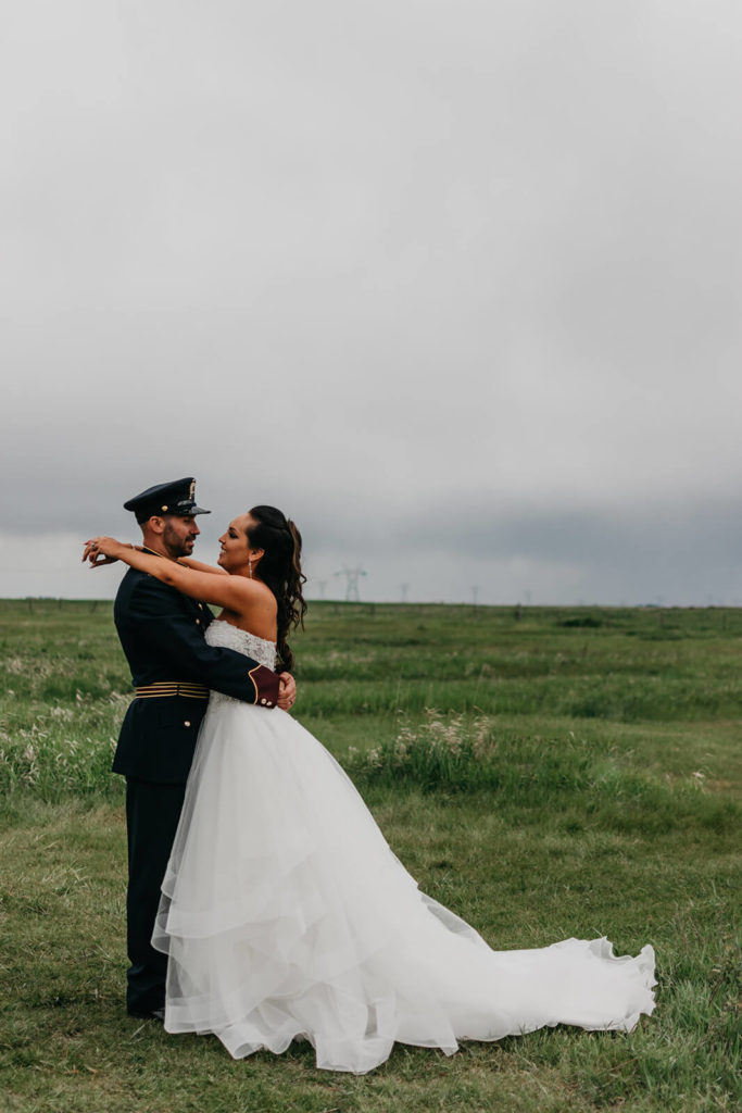 bride putting her arms around the grooms neck while they stand in a lush green field with dark grey skies