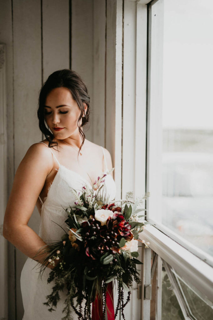 bride standing beside a window in a white cottage holding a moody flower bouquet