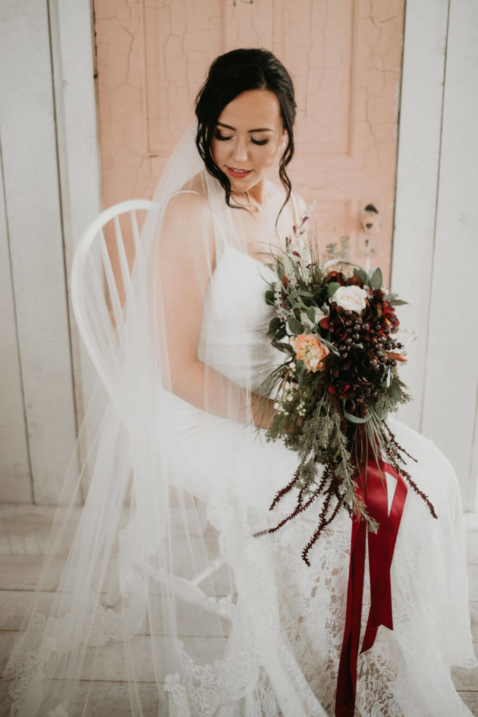 bride sitting on a chair in front of a blush pink door holding her flower bouquet