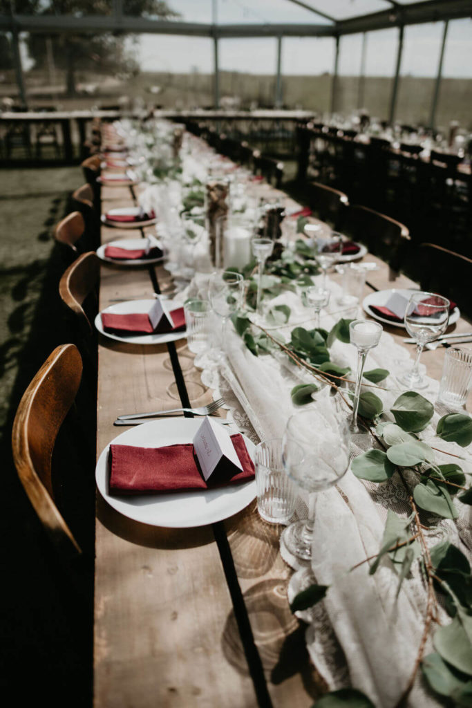 long wood tables with a white lace runner, greenery and red napkins on the plates