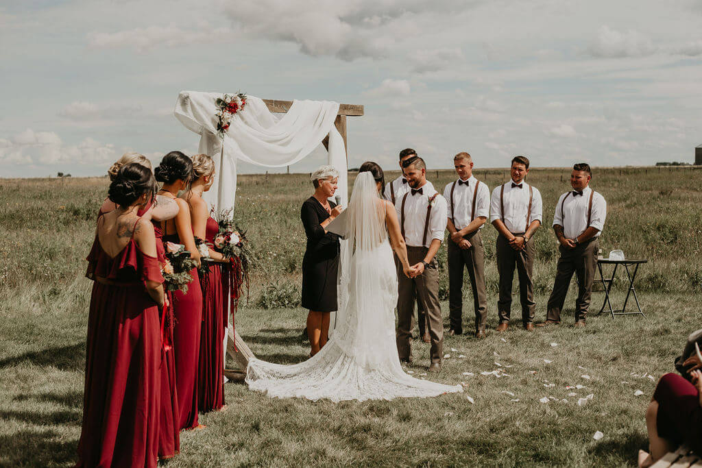 ceremony space in a open field with bridesmaids wearing red dresses and groomsmen in white shirts with leather suspenders