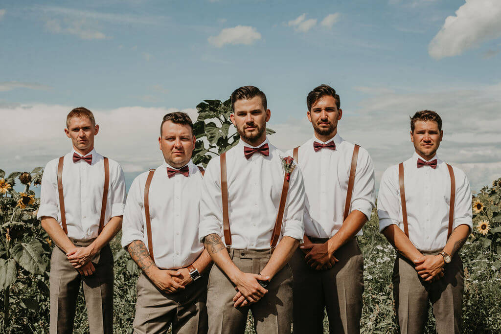 groomsmen and groom standing in a garden wearing white shirts, leather suspenders and grey pants