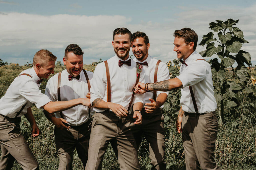 groomsmen and groom standing in a garden wearing white shirts, leather suspenders and grey pants pulling at the grooms clothes