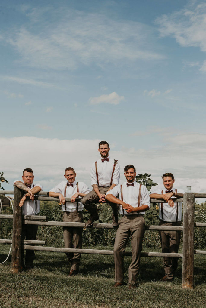 groomsmen and groom sitting on a wooden fence wearing white shirts, leather suspenders and grey pants