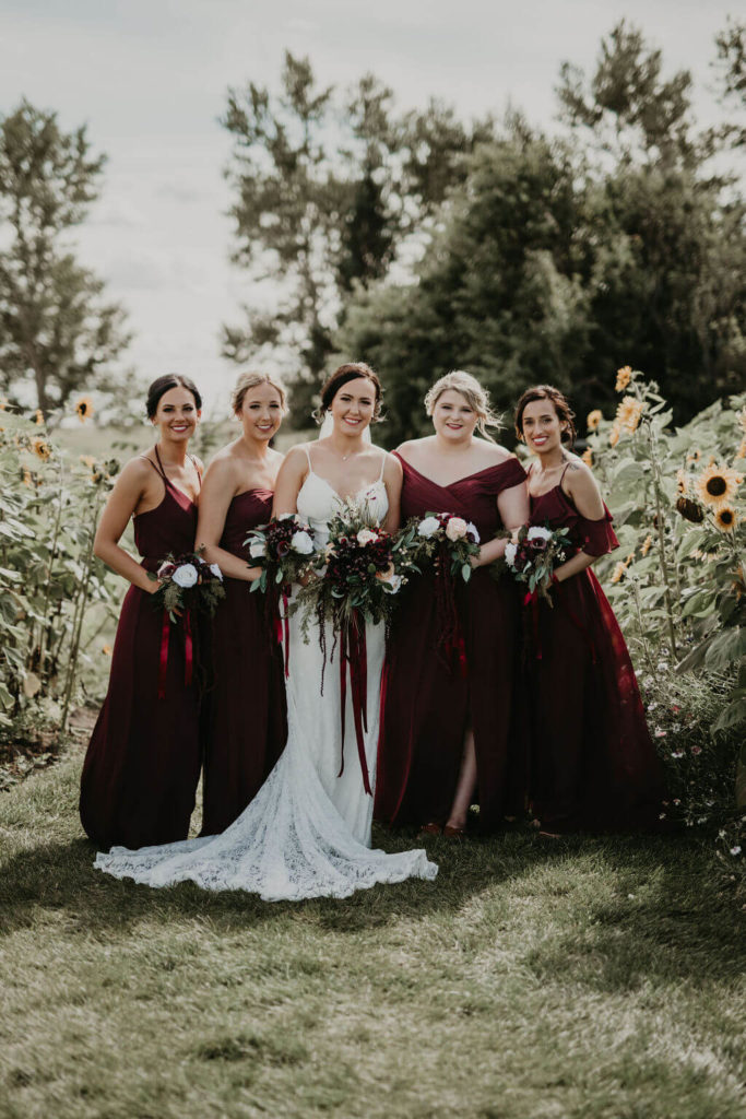 four bridesmaids wearing dark burgundy bridesmaid dresses with the bride in the middle standing in the middle of a sunflower patch