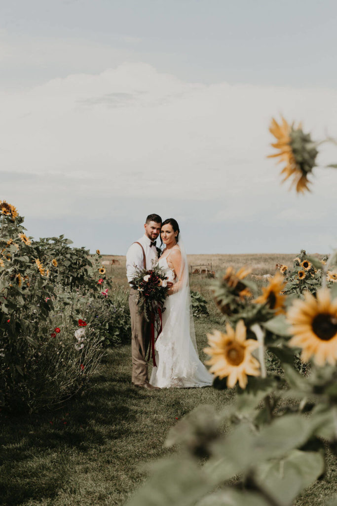 bride and groom standing in a flower field full of sunflowers, poppies and bachelor buttons