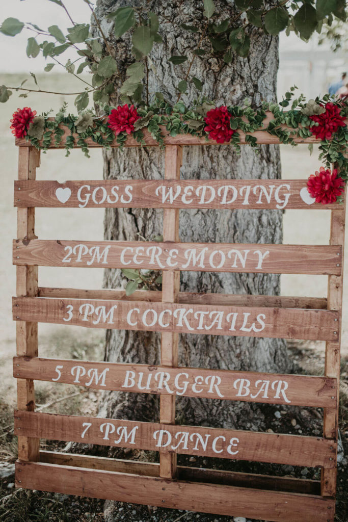 wood pallet with a wedding timeline painted on, red flowers and greenery adorn the top