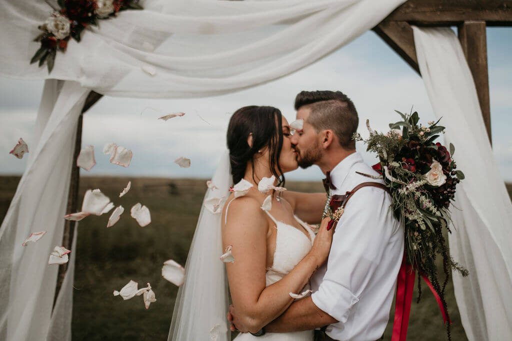 bride and groom kissing under a arbor decorated with white linen while flowers petals float around them