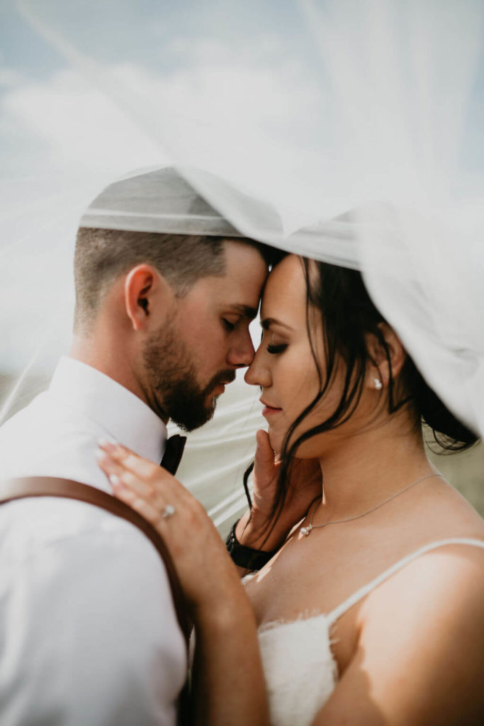 bride and groom standing with faces together while brides veil blows over them