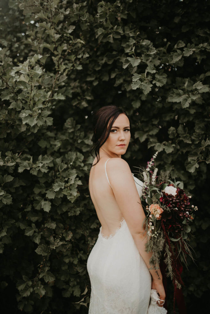 bride showing her low back dress standing in front of a green leafy tree holding her flower bouquet