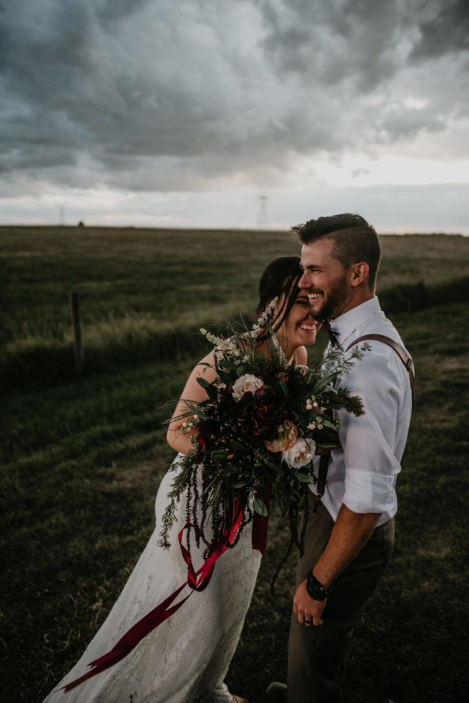 bride and groom laughing in a field while holding flowers, storm clouds above