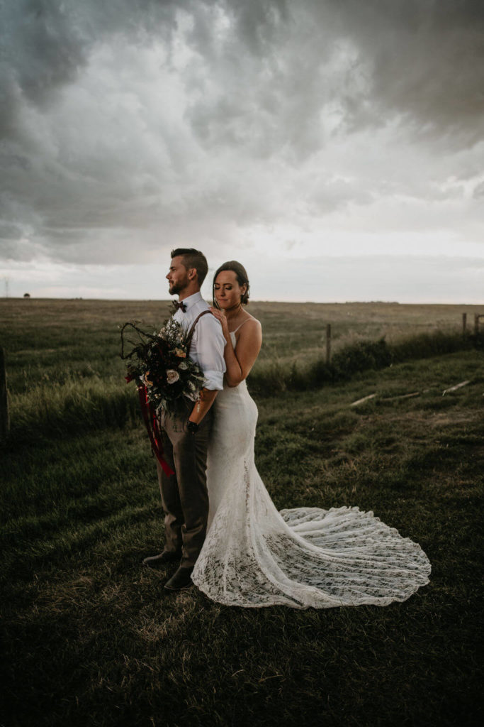 bride and groom standing in a field with a bouquet of flowers with storm clouds above