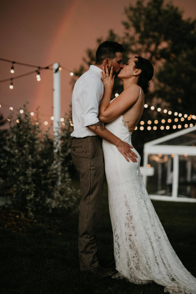 bride and groom kissing under globe lights with a pink sky and rainbow in the background