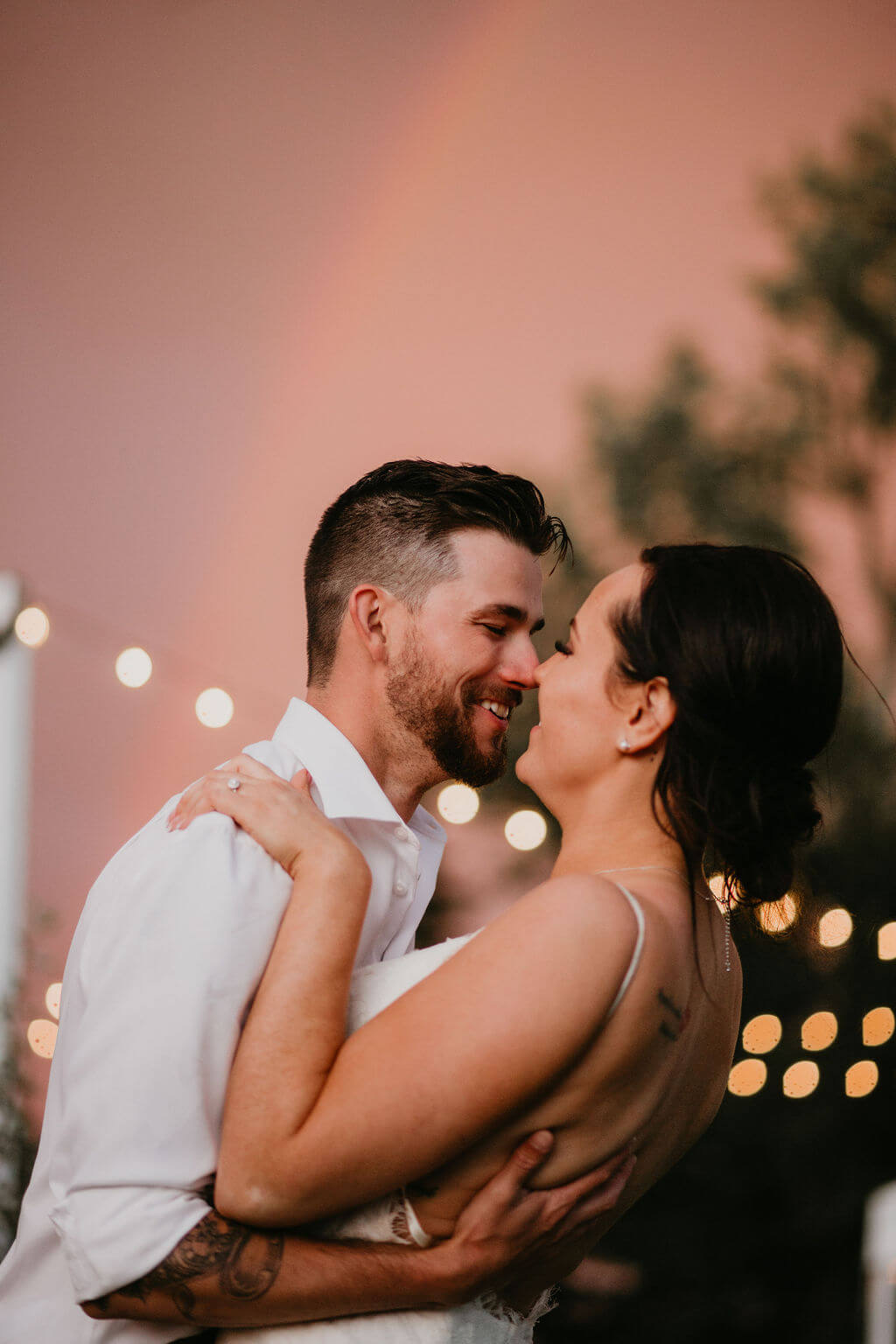 A newly married couple embrace in front of a rainbow at their outdoor wedding.