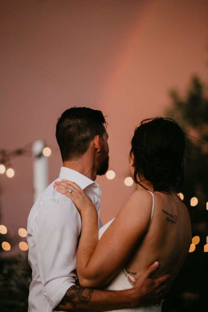 bride and groom standing under a pink sky with a rainbow