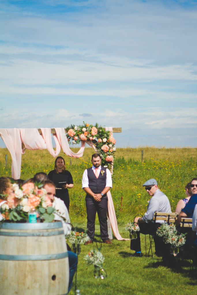 Groom in a coral tie, standing under a pink decorated wood arbor with pink and coral flowers in a prairie Alberta field.