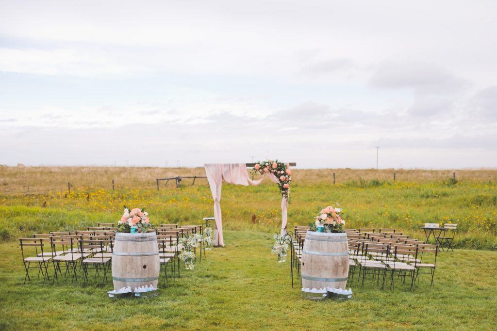 Prairie Ceremony space with wood chairs and wine barrels. Decorated with bright pinks, coral and peach tones making a vibrant appearance in the overcast sky in Alberta, Canada. The Gathered