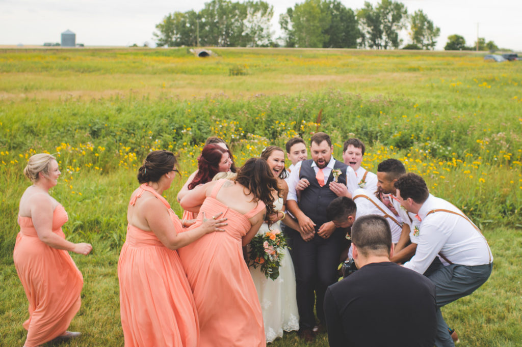 In a grassy prairie field- Bridesmaids in vibrant coral-peach flowy dresses with the groomsmen in pink bow ties at this Vibrant wedding at the Gathered.