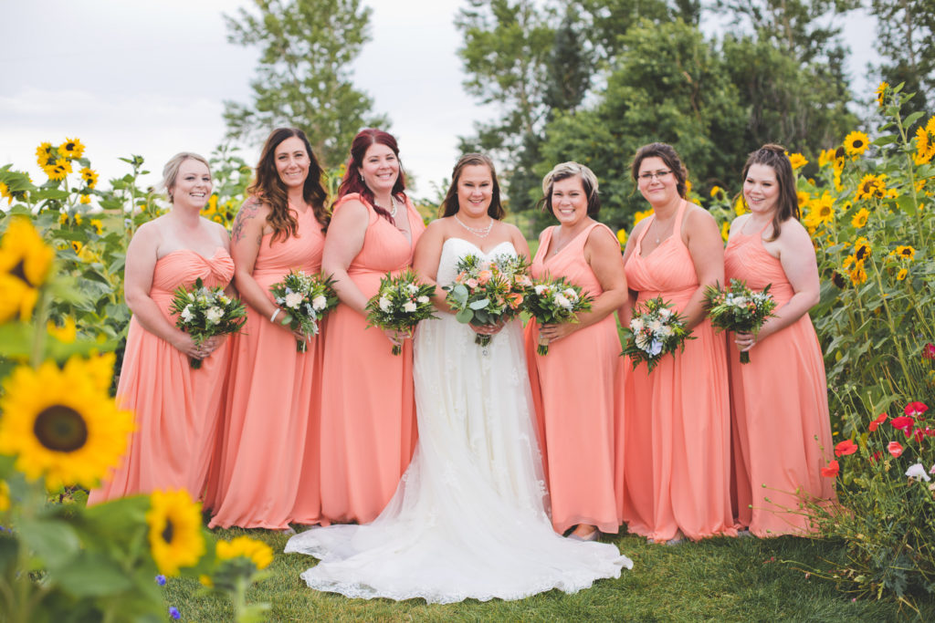 Bridesmaids in vibrant coral-peach flowy dresses stand in a sunflower field at this Vibrant wedding at the Gathered.