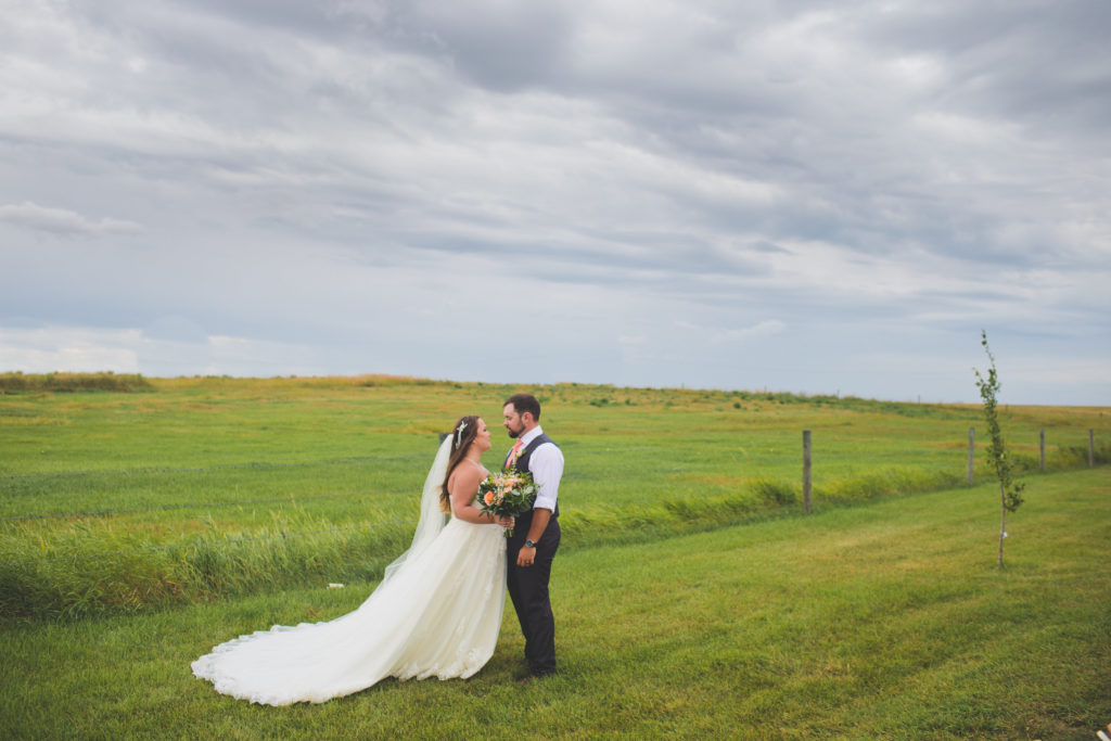 couple standing in a green prairie field with overcast skies in the background. Touches of vibrant coral and peach tones make the bouquet stand out against the brides dress on this rainy day. The Gathered in Kathyrn, Alberta