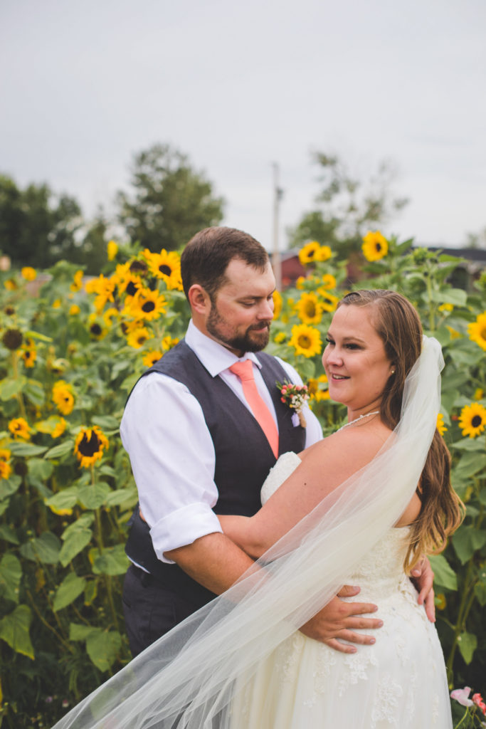 Bright yellow sunflowers surround a bride and groom as they stand in the garden. The brides veil blows in the wind and the grooms coral tie makes a pop against the sunflowers.