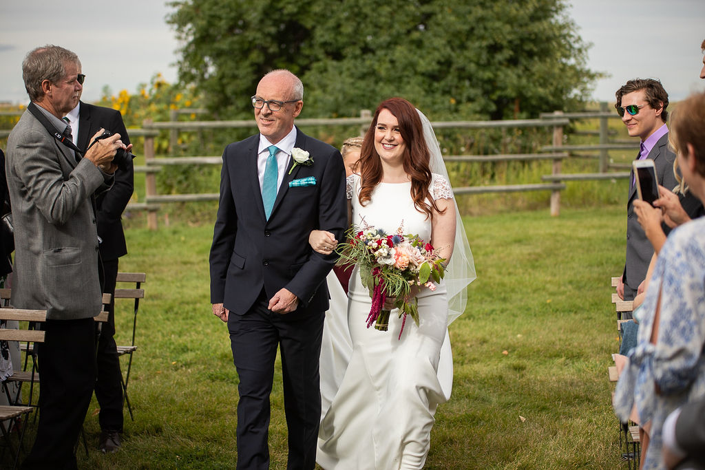 bride and father walking down a grassy floral path with wild flowers, green bushes and a farm fence surround them