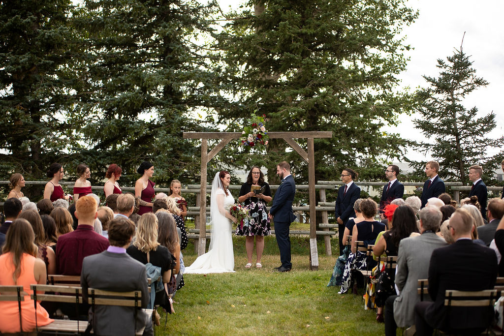 wedding couple stand under a wooden arbor with green pine trees behind them. Guests look on from their wooden chairs at this outdoor ceremony.