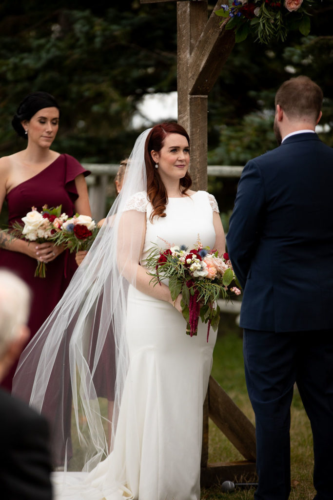 bride in a classic white dress with capped sleeves and a long veil stuns everyone with her vibrant red hair and burgundy wildflower bouquet.