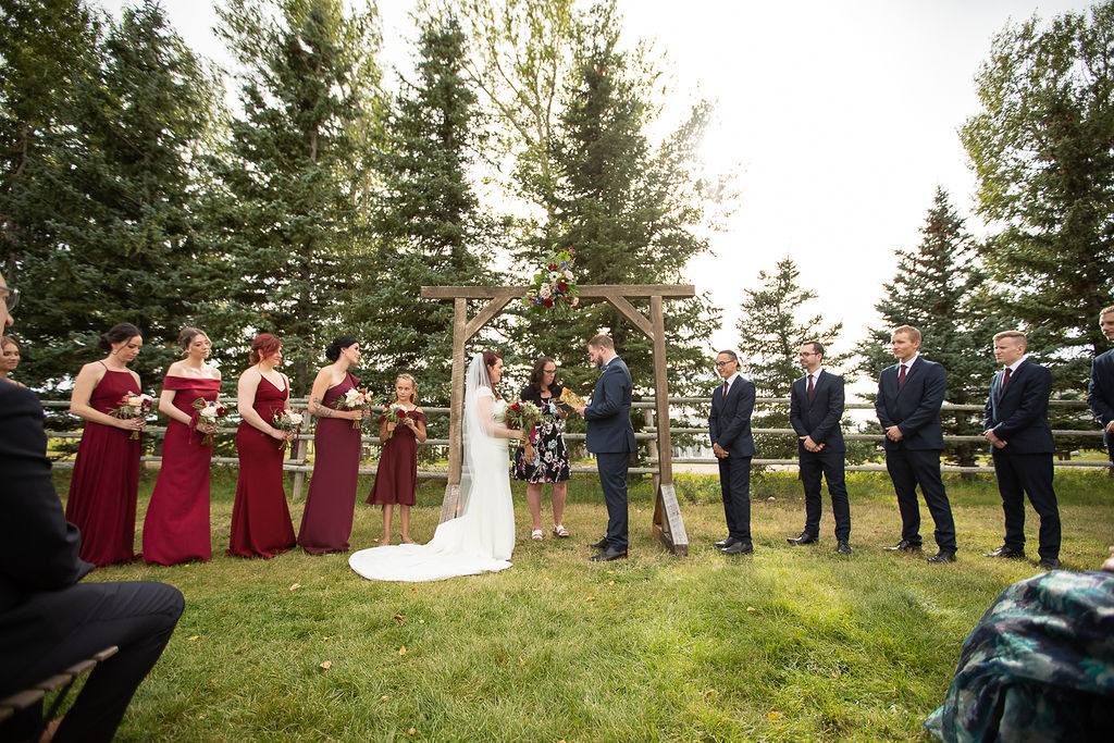 outdoor ceremony surrounded by pine trees and a wooden arbor. Bridesmaids wear long burgundy dresses and the groomsmen have burgundy ties.