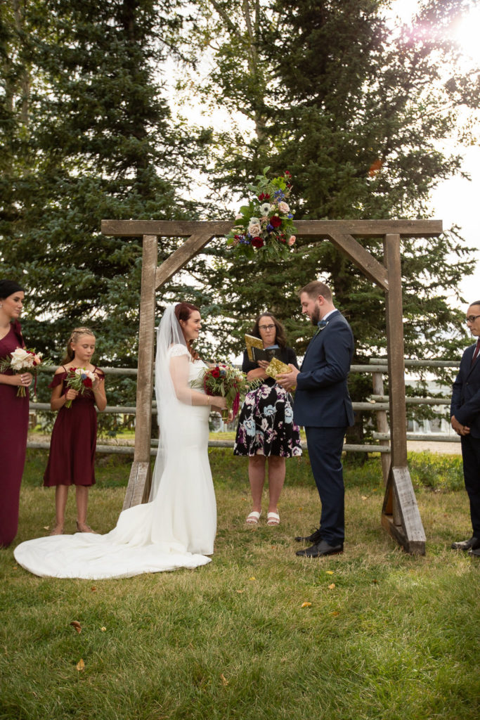 outdoor ceremony surrounded by pine trees and a wooden arbor. Bridesmaids wear long burgundy dresses and the groomsmen have burgundy ties.