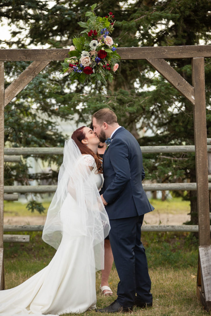 just married kiss under a wooden arbor at this outdoor ceremony space - surrounded by big green trees and loving guests .
