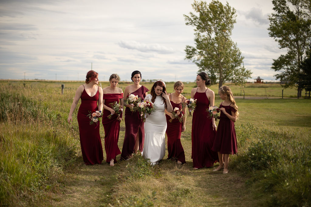 6 bridesmaids in burgundy floor length dresses surround a bride while standing in a prairie field. www.thegathered.ca