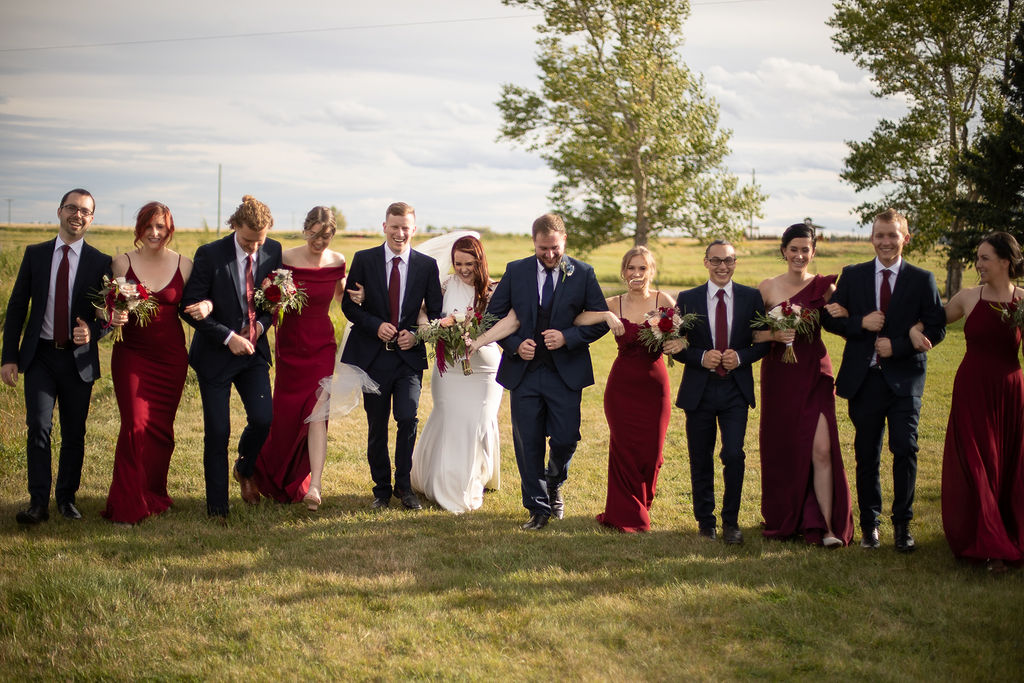 bridal party dresses in burgundy tones surround the bride and groom as they all walked with arms linked through a field. www.thegathered.ca