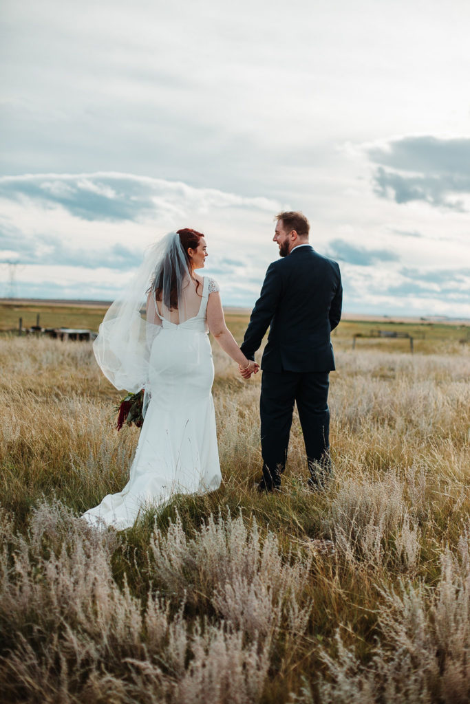 bride and groom walk through a wild overgrown prairie field with an cloudy sky in the background. www.thegathered.ca