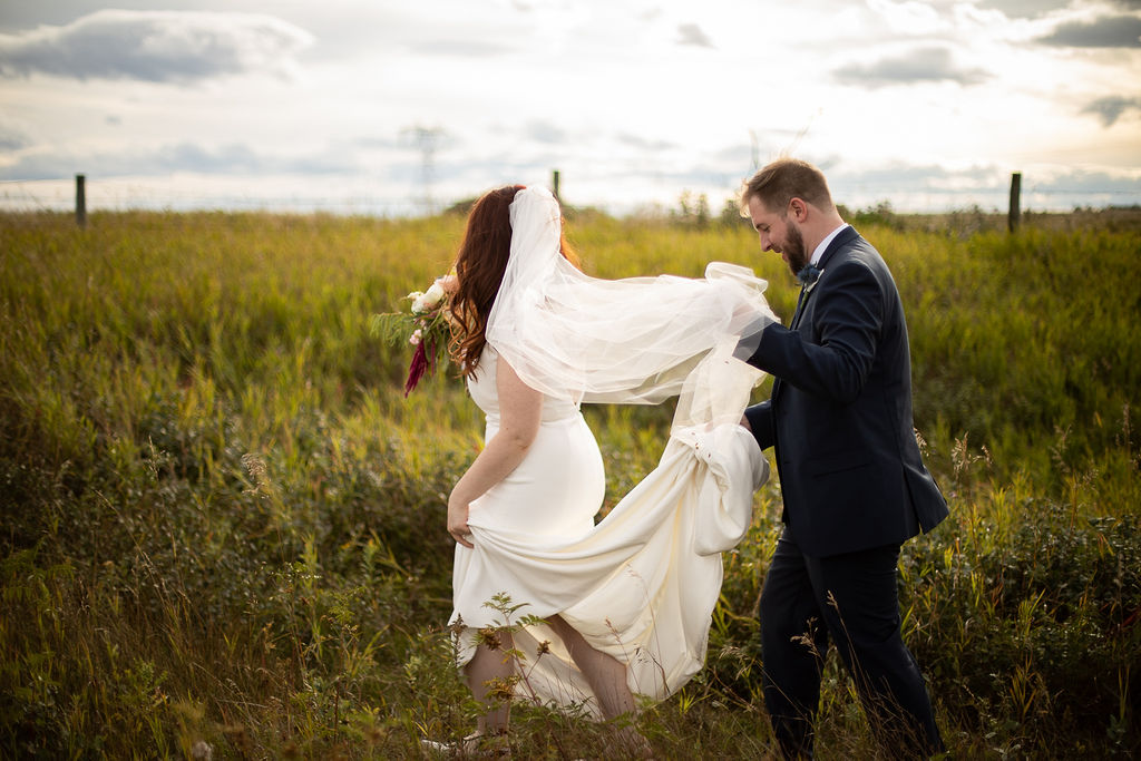 a couple walks through wild prairie fields - the groom helps the bride with her long dress train and her veil. the sun sets in the back. www.thegathered.ca