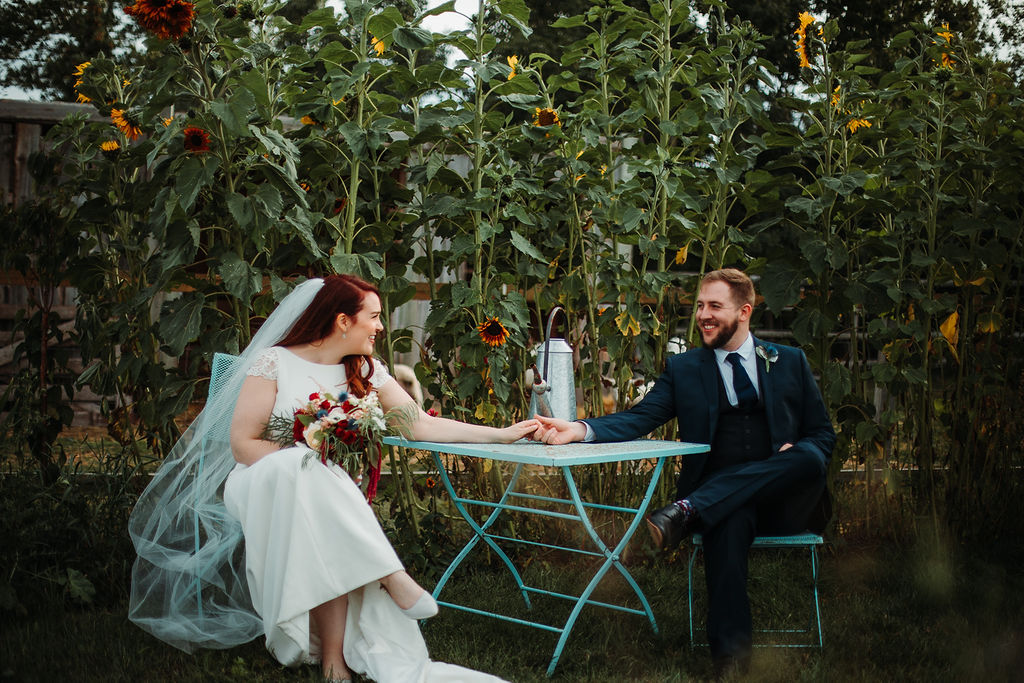 bride and groom sit at a blue table holding hands across the table as a wall of giant sunflowers make a backdrop behind them. www.thegathered.ca