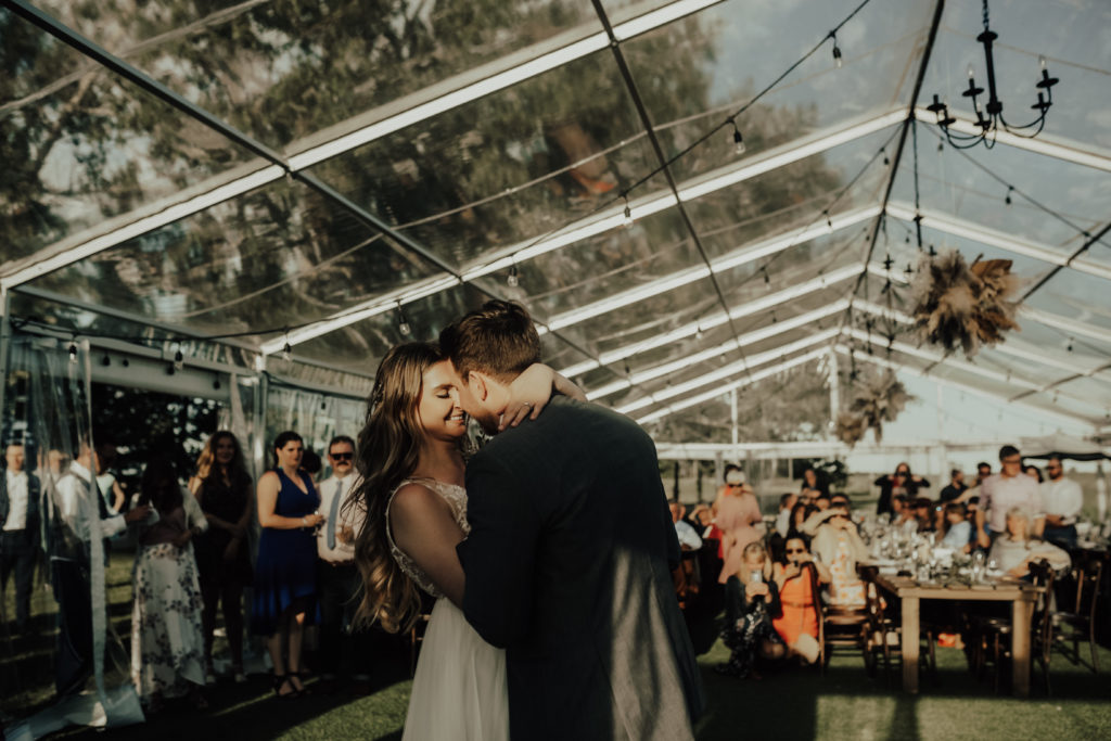 couple enjoying their first dance in a clear tent with wedding guests mingling around them as the sunsets