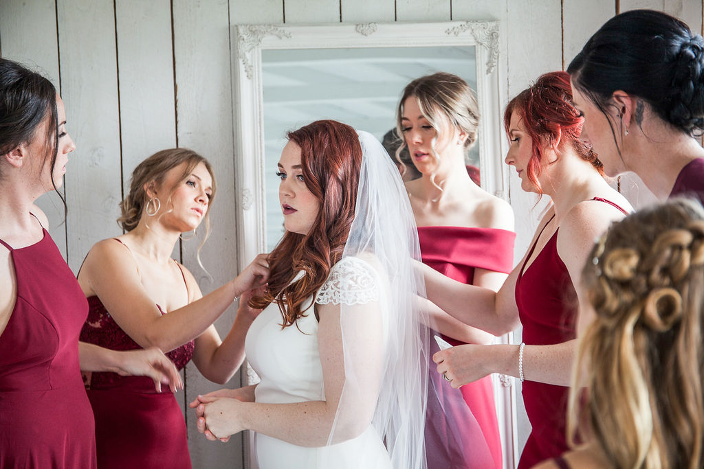 bridesmaids in burgundy wedding dresses help a bride with her veil