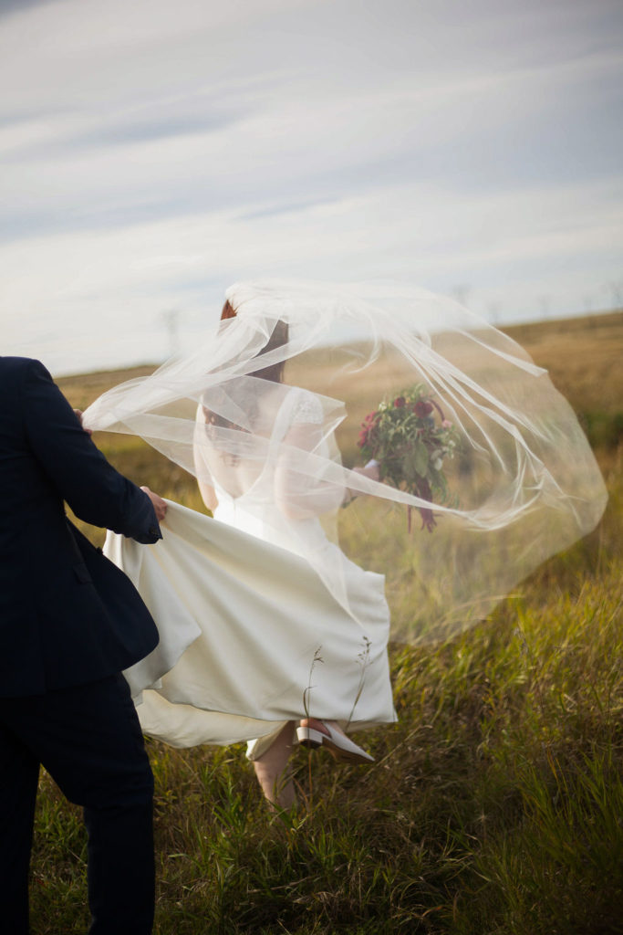 brides veil blows wildly in the wind as they walk as she walks through a field. www.thegathered.ca