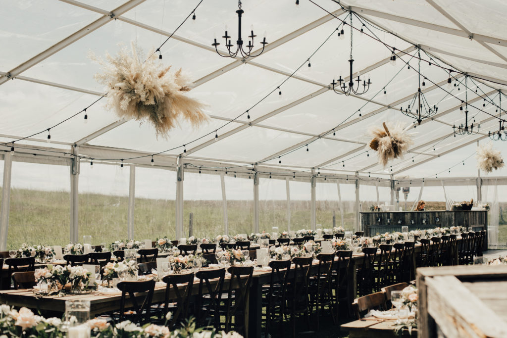 clear tent with hanging chandeliers and globe lights, pampas hanging floral pieces with long rows of wood tables a plenty of flower centrepieces. www.thegathered.ca