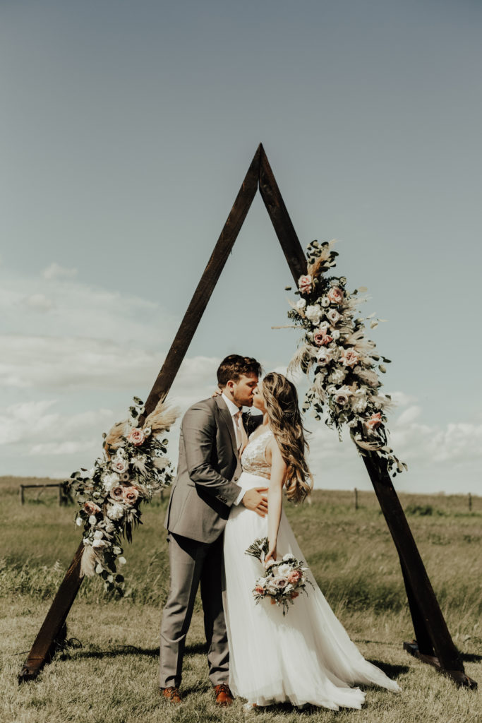 bride and groom kiss under a triangle arbor with floral arrangements adorning the arch. Arbor is standing in a prairie field. www.thegathered.ca