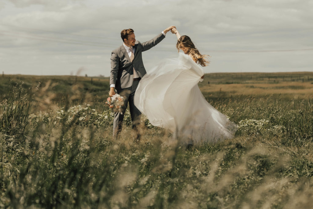 groom twirls the bride in a circle while her dress blows in the wind as they stand in a grassy prairie field. www.thegathered.ca