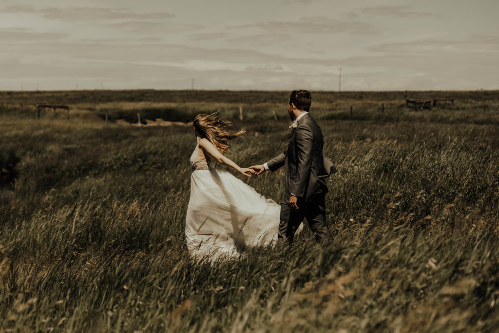 groom walks behind the bride holding her hand while her dress blows in the wind as they stand in a grassy prairie field. www.thegathered.ca