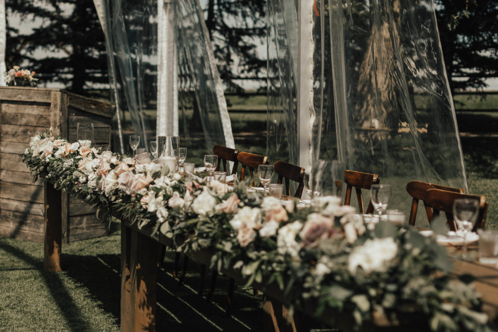 head table lined completely with white and blush roses with greenery mixed in. Outdoor wedding venue in Calgary Alberta.