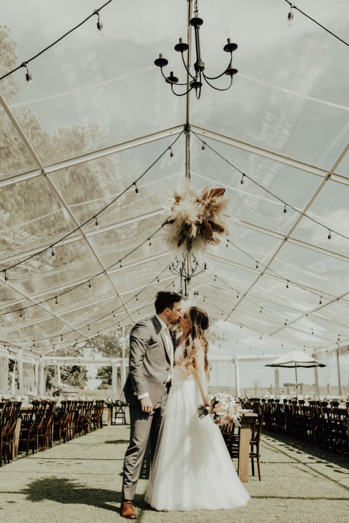 bride and groom kiss in their clear wedding reception tent. The hanging globe lights, black chandeliers and pampas grass hanging piece add the perfect amount of modern flair. www.thegathered.ca